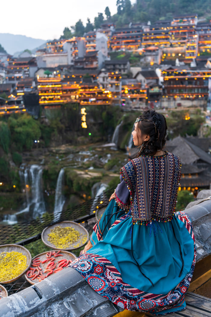 Mujer con vestimenta tradicional contemplando la vista panorámica de la ciudad antigua de Lijiang iluminada por la noche junto a una cascada.