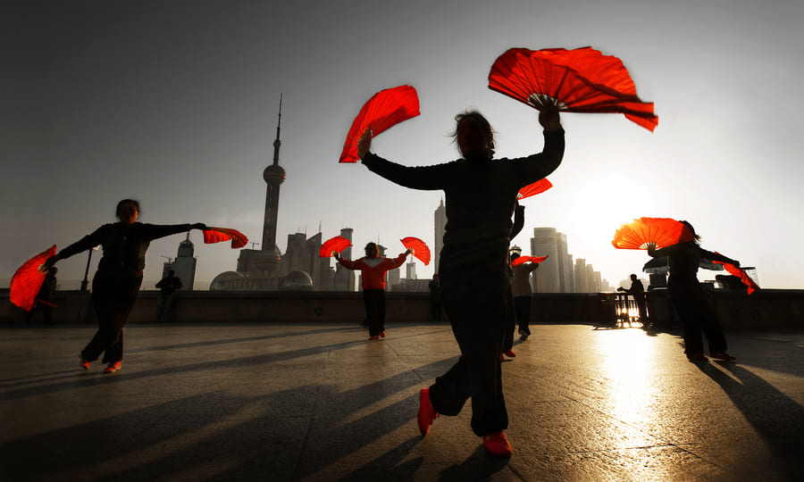Personas practicando danza tradicional con abanicos rojos en el Bund de Shanghái, con el horizonte de Pudong de fondo al amanecer.