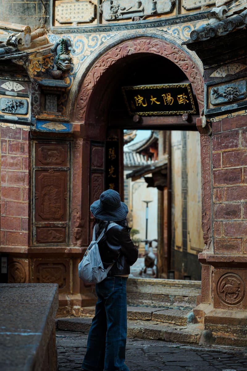 Una turista con sombrero de pescador y mochila observa desde fuera un patio antiguo a través del arco de piedra tallada de una edificación histórica china.